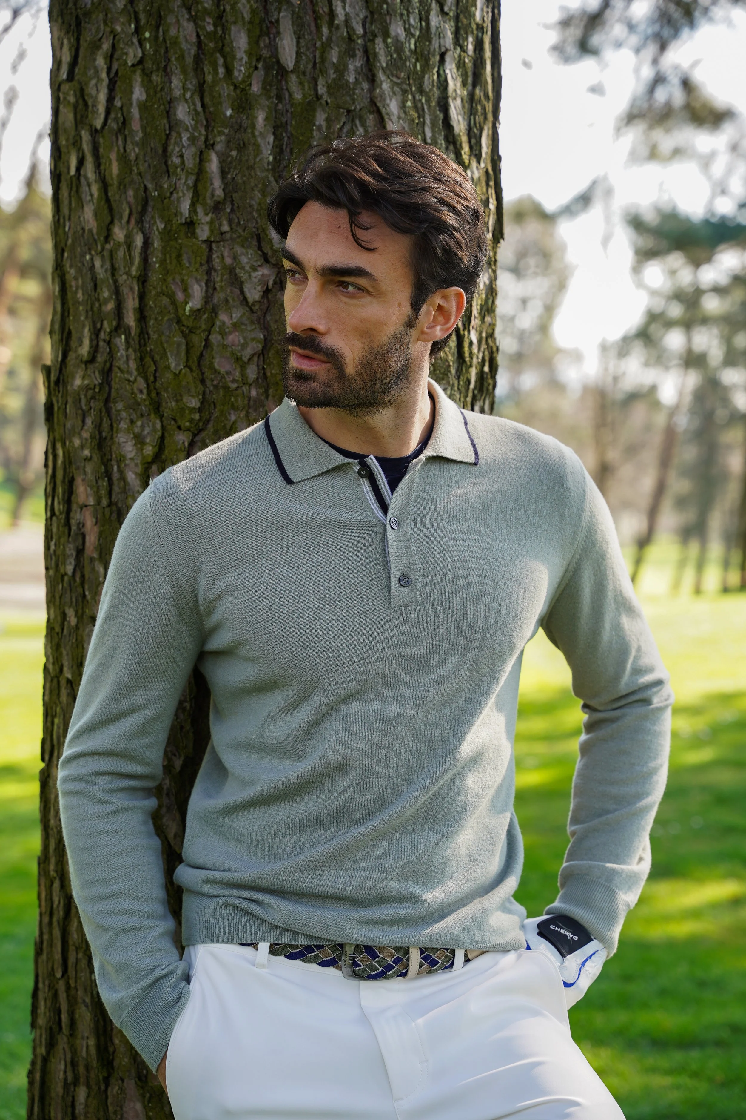Honey+Haze Photography. A man with dark hair and facial hair leaning against a tree outdoors, wearing a light gray long-sleeve polo shirt and white pants with a patterned belt, in a park with green grass and trees in the background.