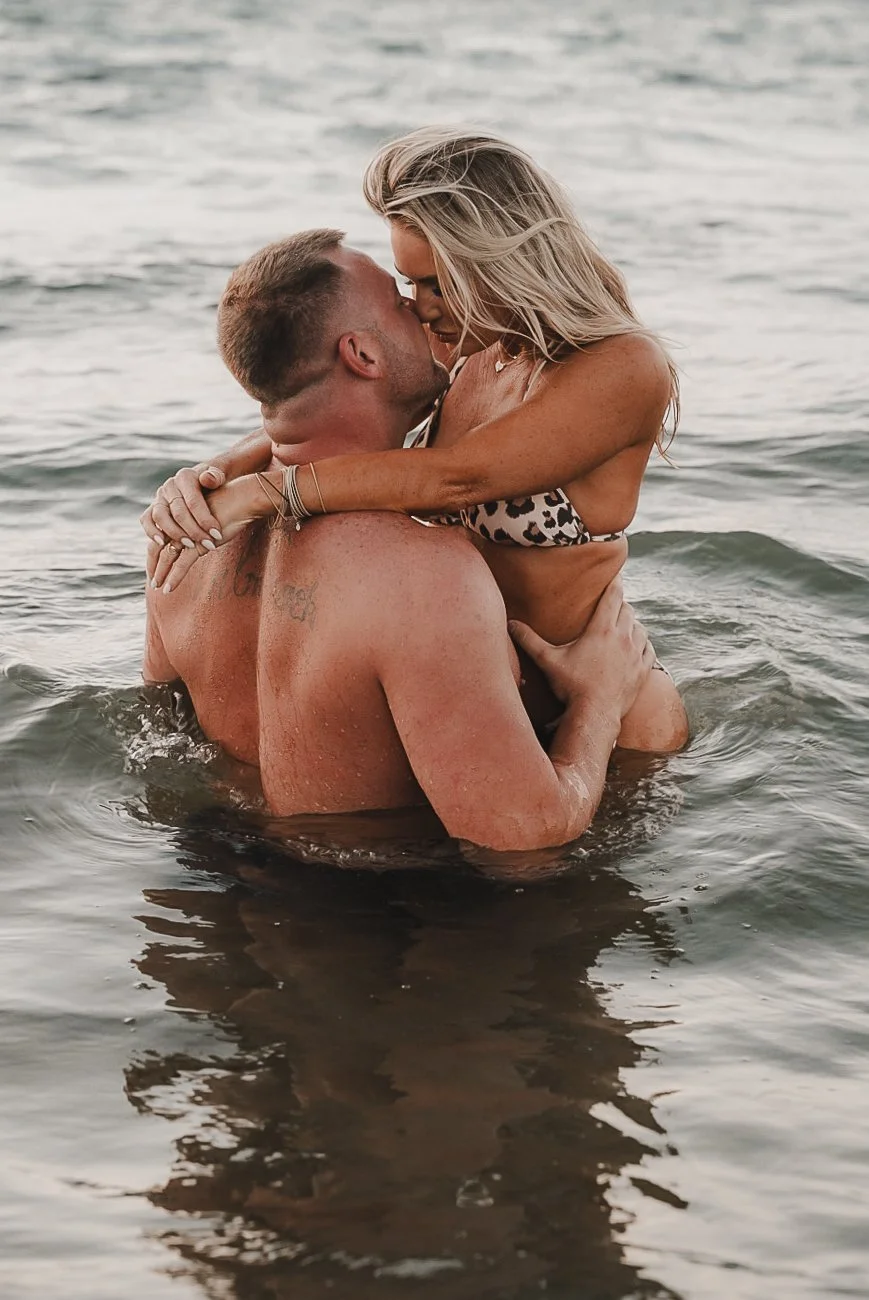 Honey+Haze Photography. A man and woman are embracing and kissing in the water, with the woman sitting on the man's shoulders.