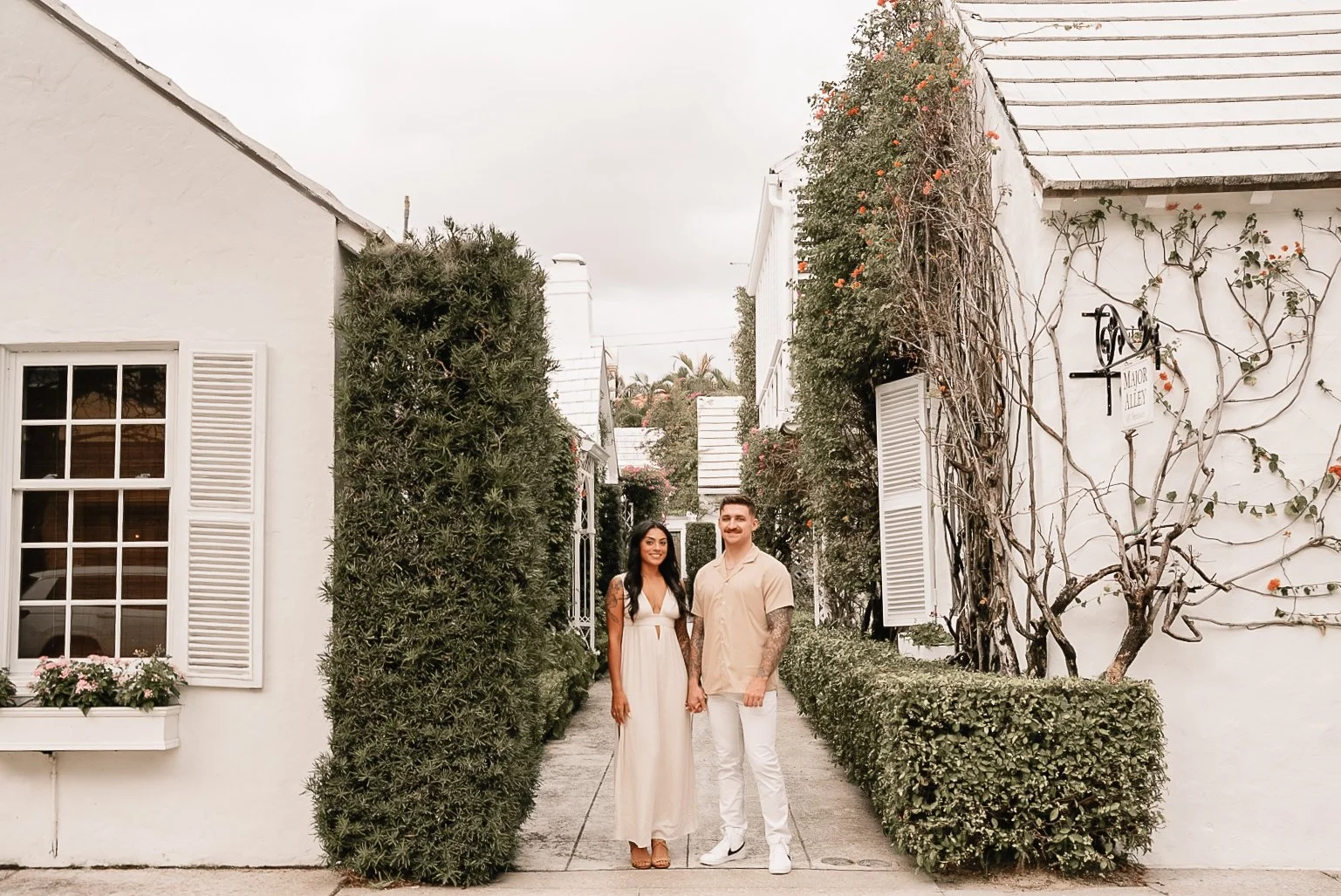 Honey+Haze Photography. A man and woman holding hands on a sidewalk, with white buildings, trimmed hedges, and flowering vines in the background.