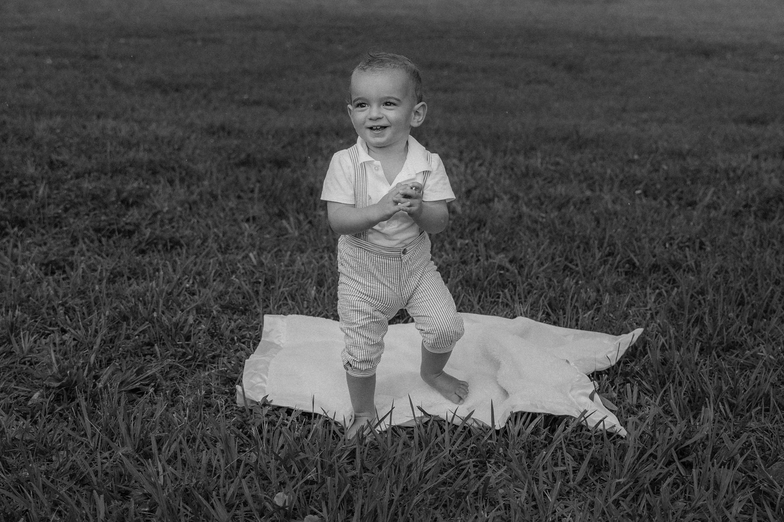 Honey+Haze Photography. A young boy is standing on a blanket outdoors on grass, smiling and holding his hands together, with more grass and a blurred background in black and white.