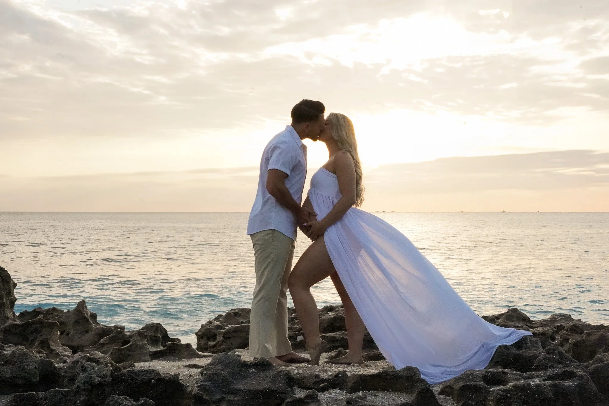 Honey+Haze Photography. A couple kissing on a rocky beach during sunset, with water and the sky in the background.