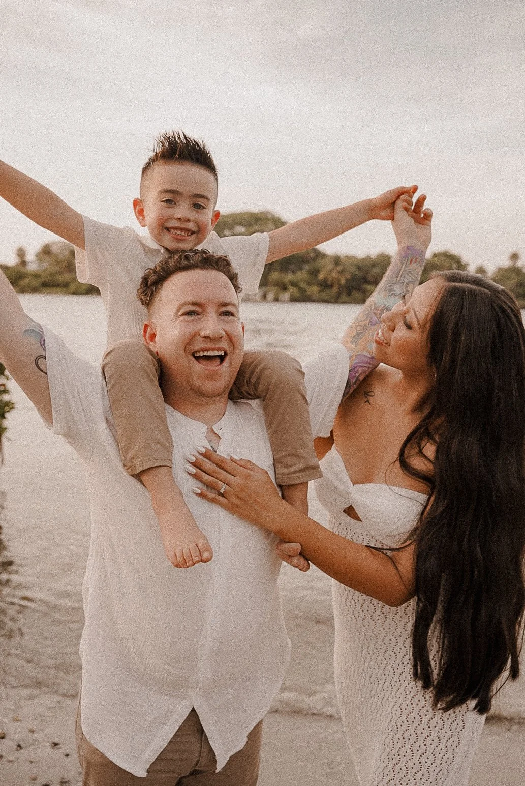 Honey+Haze Photography. Family of three enjoying time together at the beach, with a man giving a piggyback ride to a boy, and a woman smiling at them, with water and trees in the background.