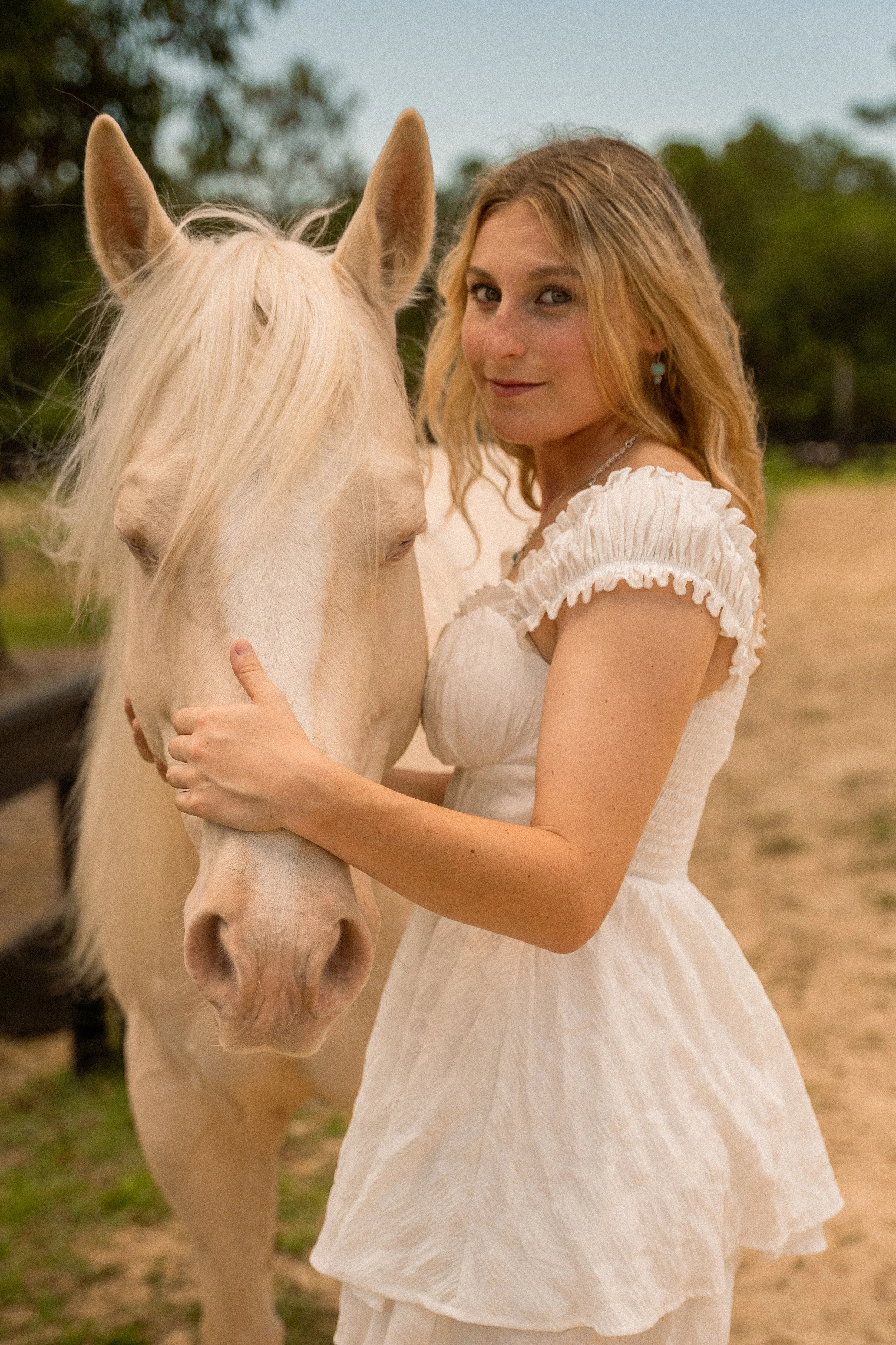 Honey+Haze Photography. A woman with long, wavy, red hair wearing a white dress with ruffled sleeves, standing outdoors and holding a white horse's face close to her.