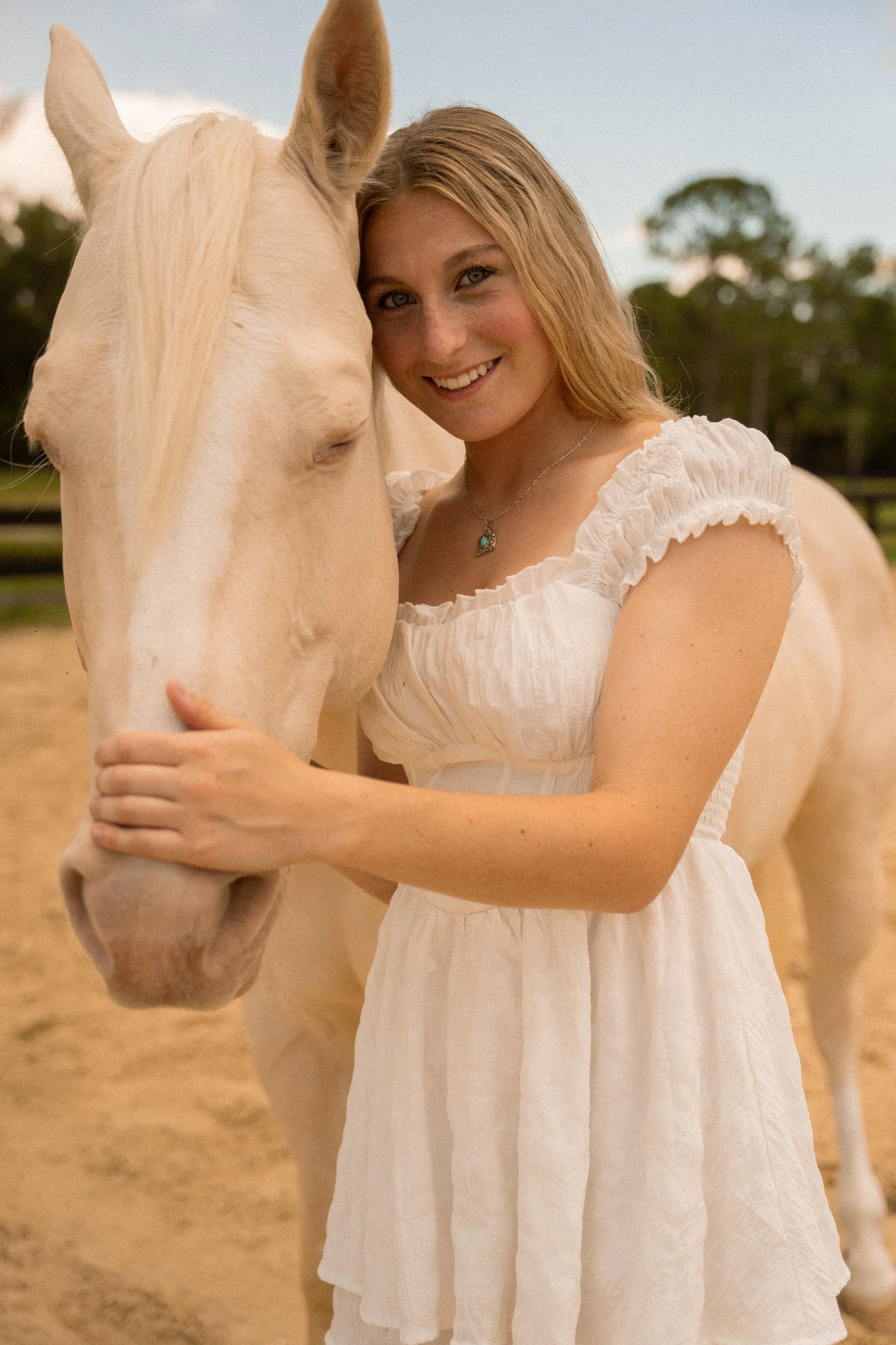 Honey+Haze Photography. A woman with blonde hair wearing a white dress is smiling and hugging a light-colored horse in an outdoor setting with trees and a fence in the background.