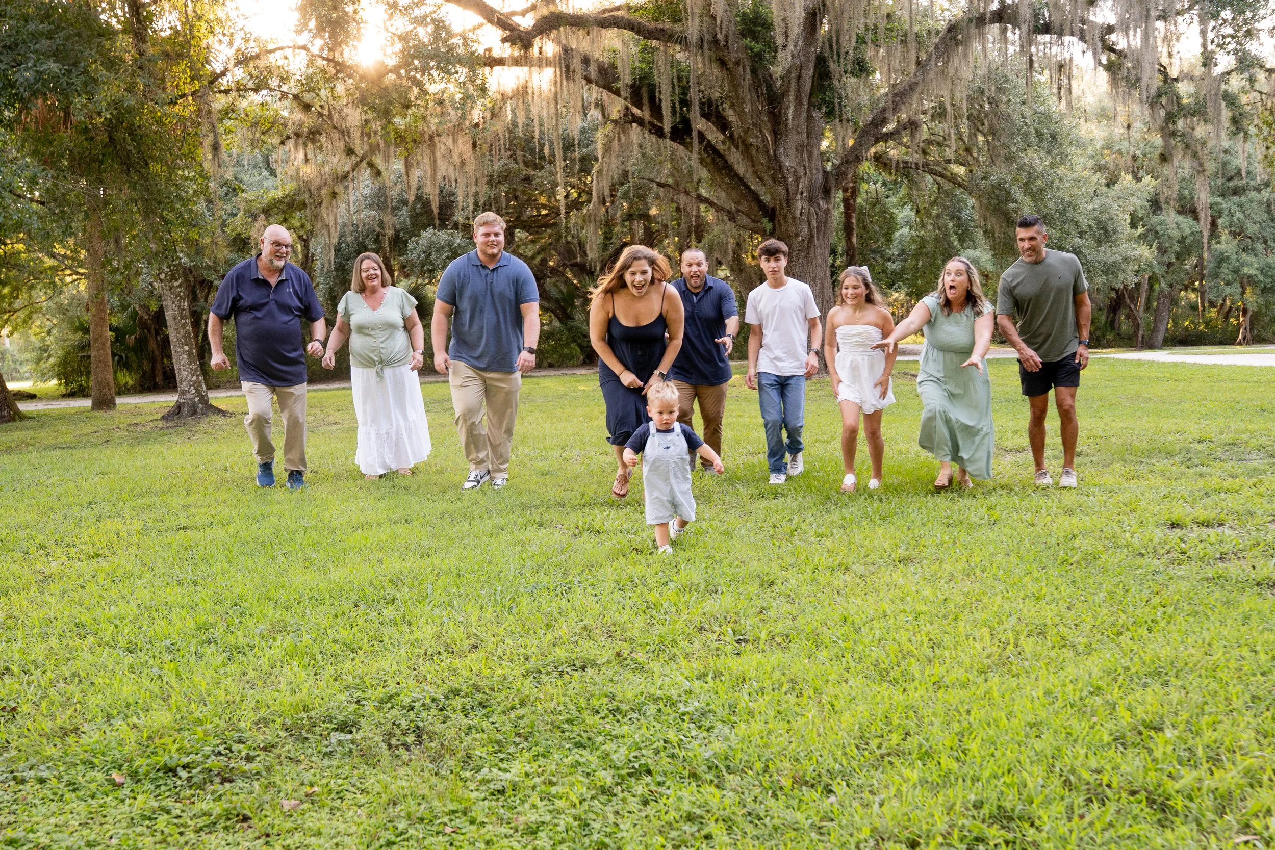 Honey+Haze Photography. A group of adults and children smiling and walking together on a grassy field, with large trees and Spanish moss hanging from the branches in the background, during the golden hour sunlight.