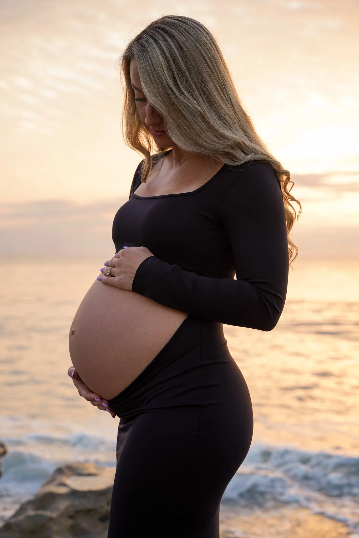 Honey+Haze Photography. A pregnant woman in a black dress standing on the beach at sunset, looking down at her belly, with her hands gently resting on her abdomen.