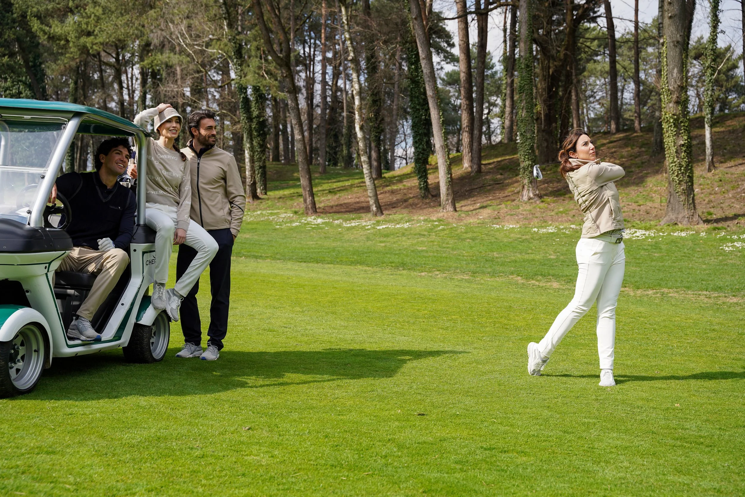Honey+Haze Photography. A woman in white pants and a beige jacket swings a golf club on a golf course, while three friends watch from a golf cart nearby among trees.