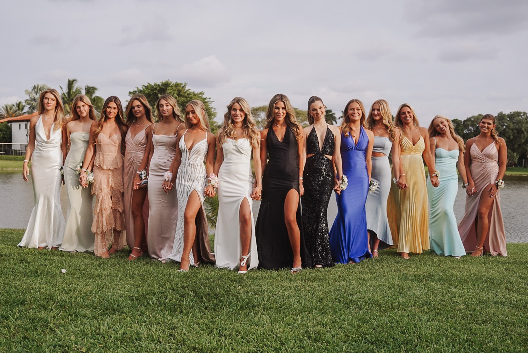 Honey+Haze Photography. Group of women in colorful evening gowns standing outdoors on grass, with trees and cloudy sky in the background.