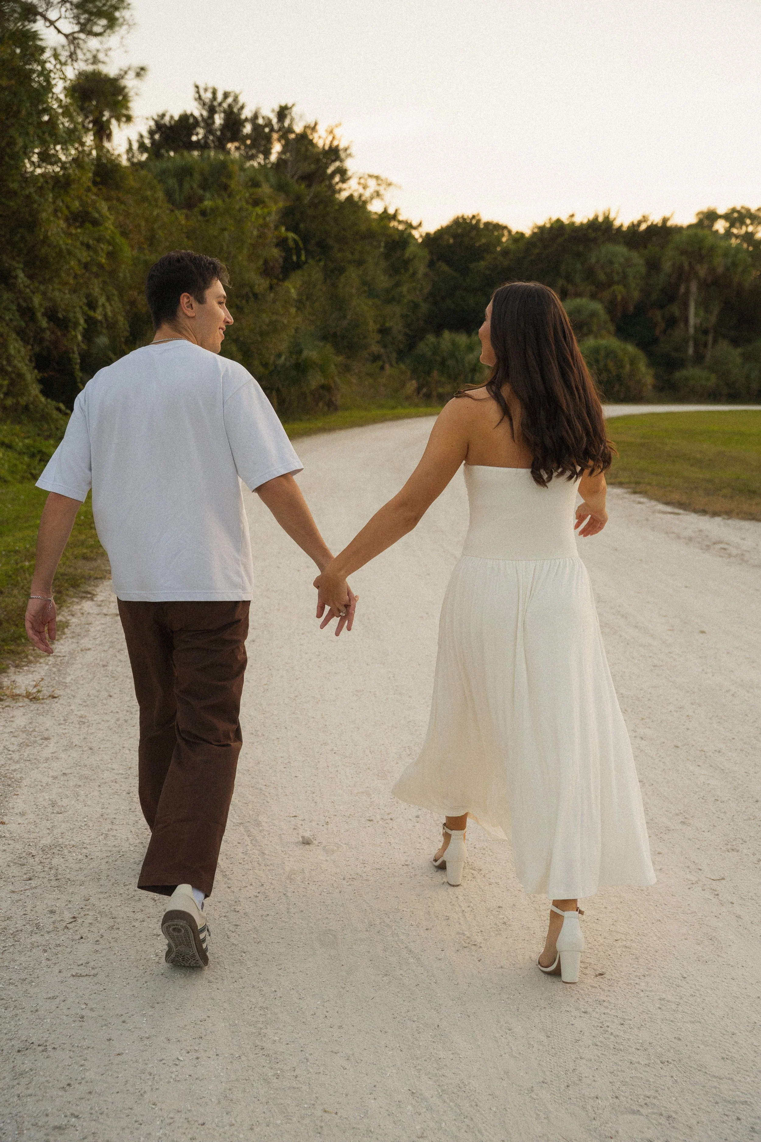 Honey+Haze Photography. A young couple walking hand in hand on a gravel path surrounded by greenery, with the sun setting in the background. The woman wears a white dress and heels, and the man wears a white shirt and brown pants.