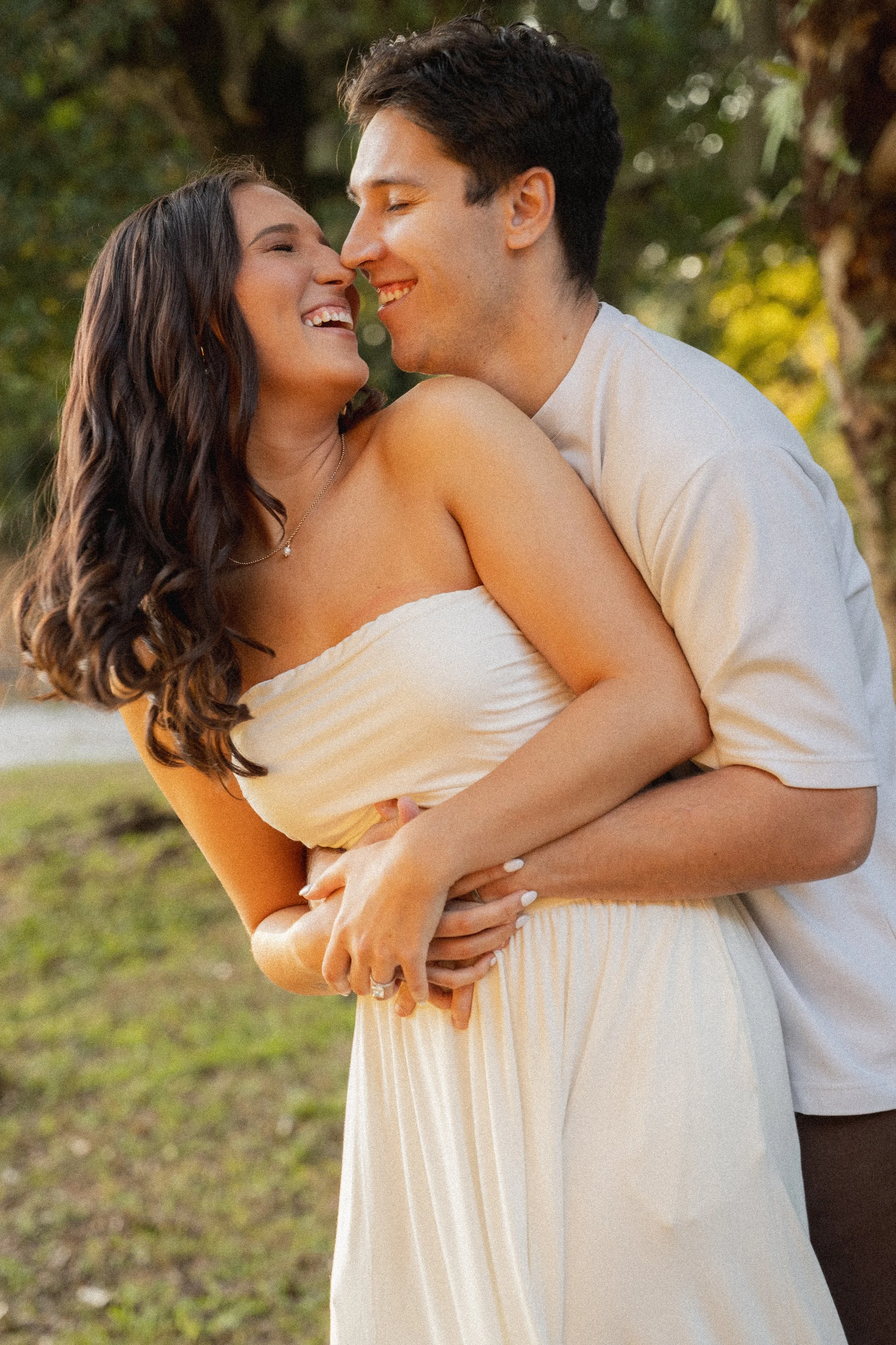 Honey+Haze Photography. A couple is embracing outdoors, smiling and with their noses touching. The woman is wearing a strapless yellow dress and a pearl necklace, and the man is in a white shirt. They are surrounded by trees and sunlight.