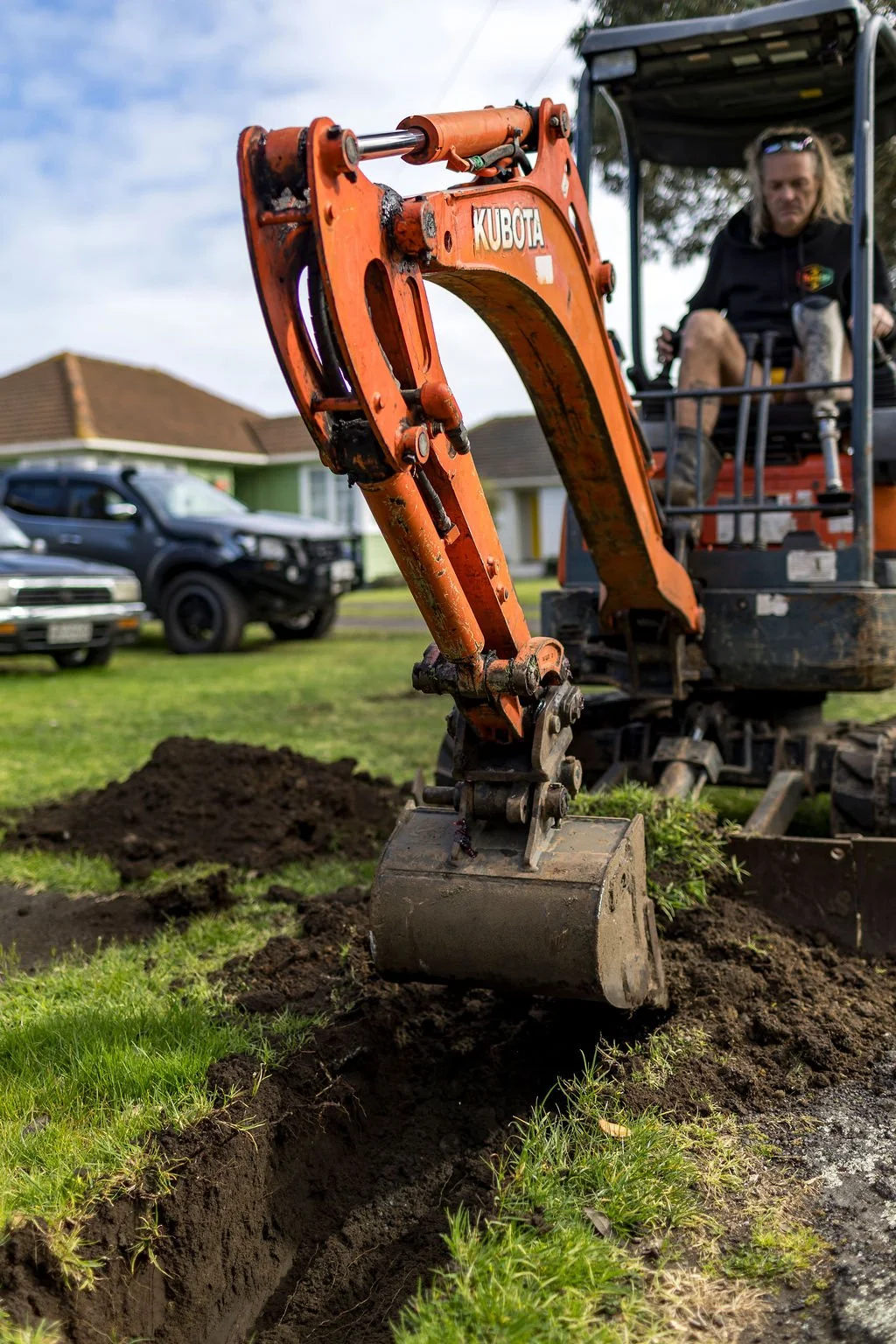 A worker operating an orange Kubota mini excavator digging a trench in a grassy yard. Several parked cars and houses are visible in the background.