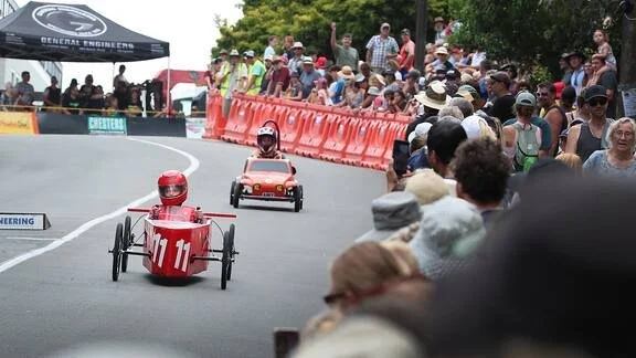 Children racing small go-karts on a street with spectators watching from both sides.