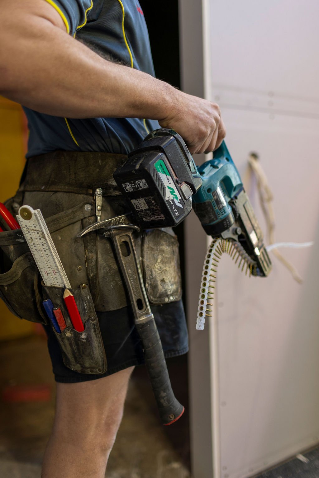 Close-up of a worker's waist and hands holding power tools, including a cordless drill and a nail gun, with various tools in a tool belt.