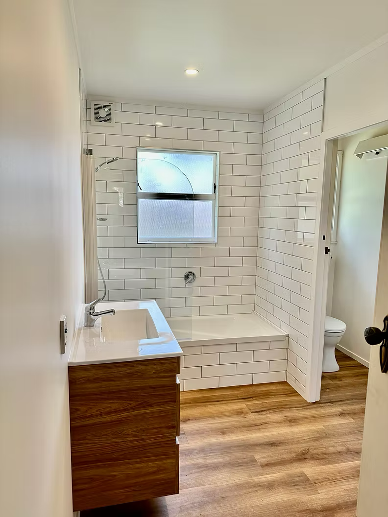 A small bathroom with a white tiled shower area, a frosted window, a wooden vanity with a white sink, an overhead light, and a toilet visible in an adjacent room.