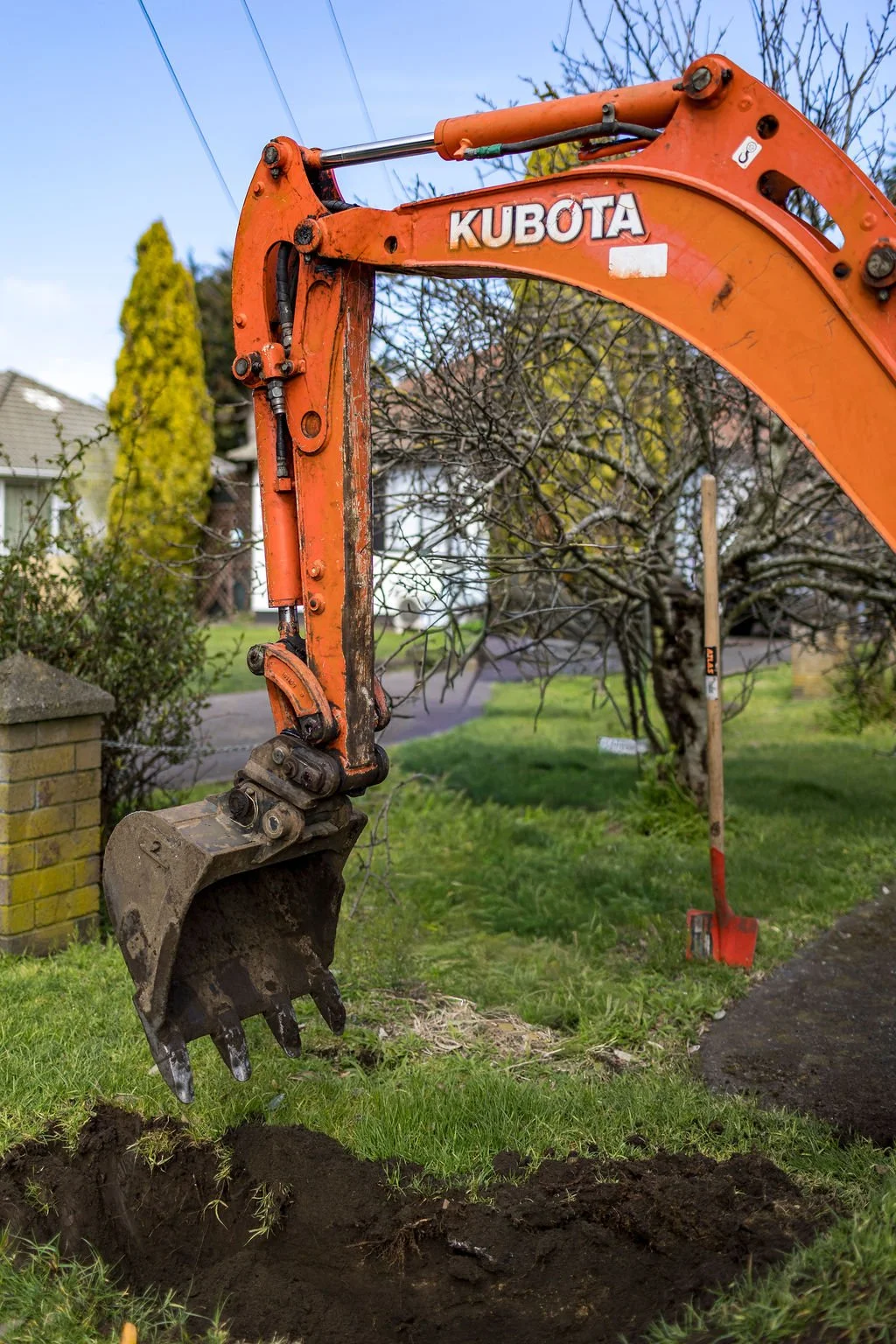 An orange Kubota mini excavator digging a hole in a grassy yard with trees and houses in the background.