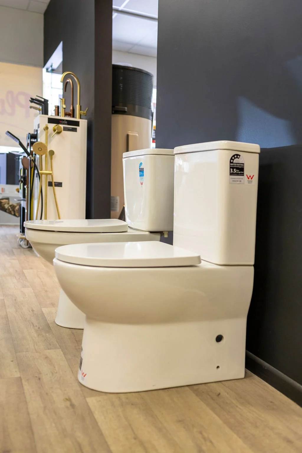 Two white toilets placed side by side in a showroom or store display, with other bathroom fixtures visible in the background.