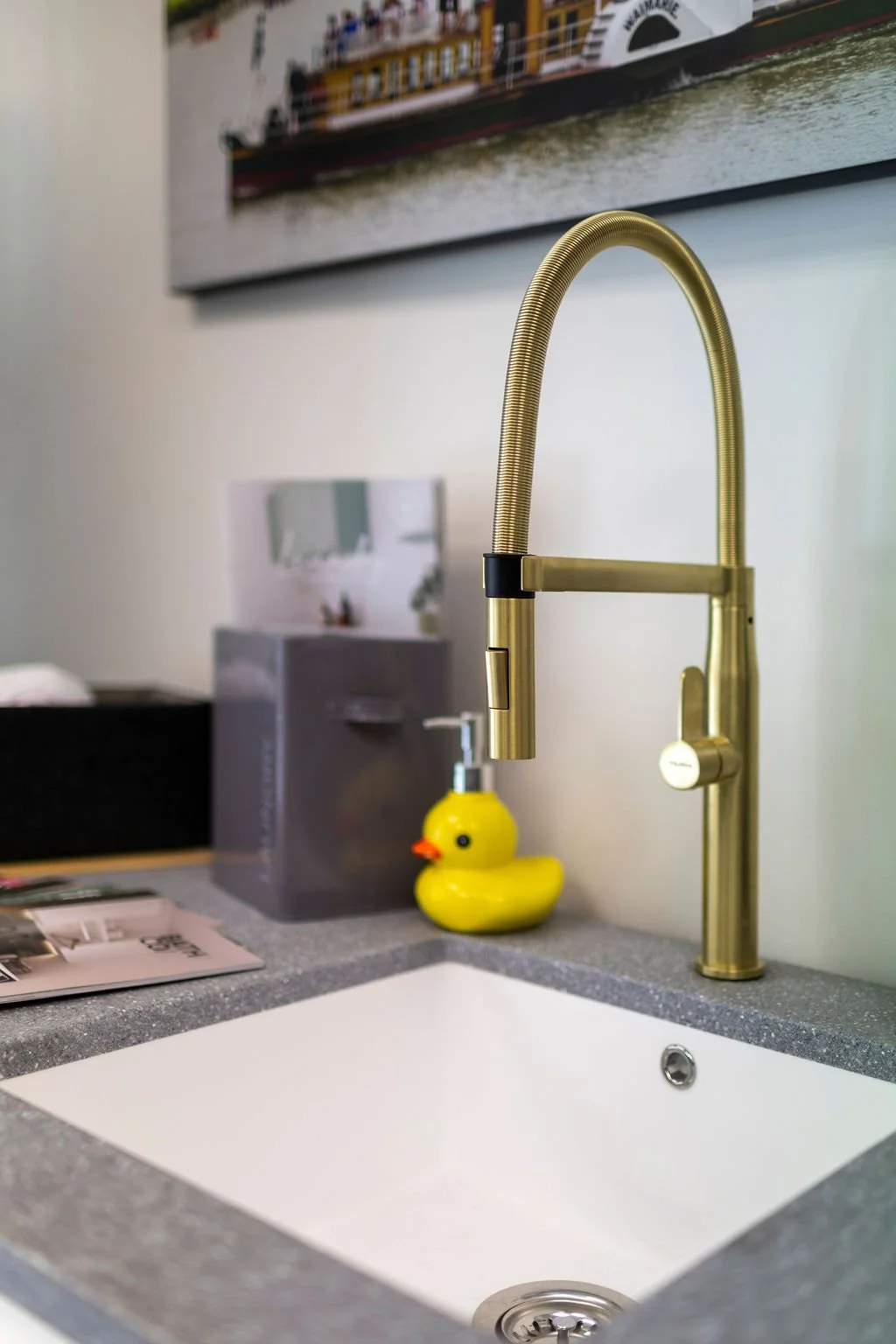 Close-up of a gold kitchen faucet with a spring coil design on the neck, mounted above a white sink with a gray countertop. A yellow rubber duck soap dispenser is on the counter, with a box and a magazine nearby. A blurred picture of a ship displays on the wall in the background.