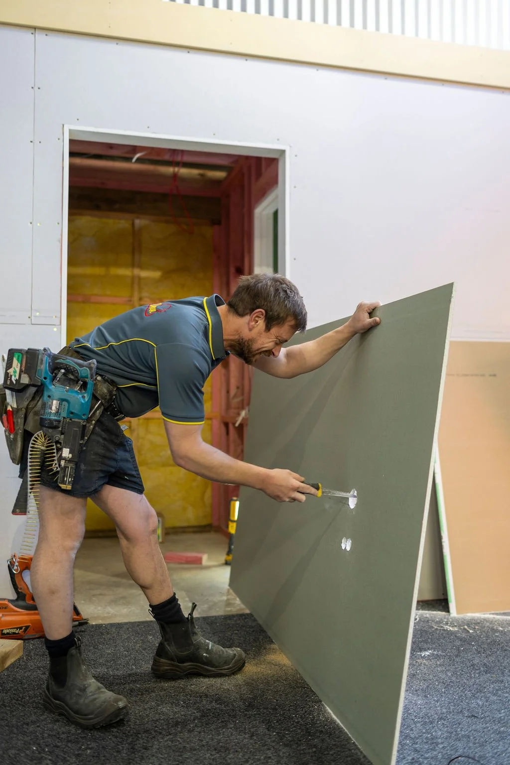 A man working on drywall inside a building under construction, holding a screwdriver and posing with a large sheet of drywall.