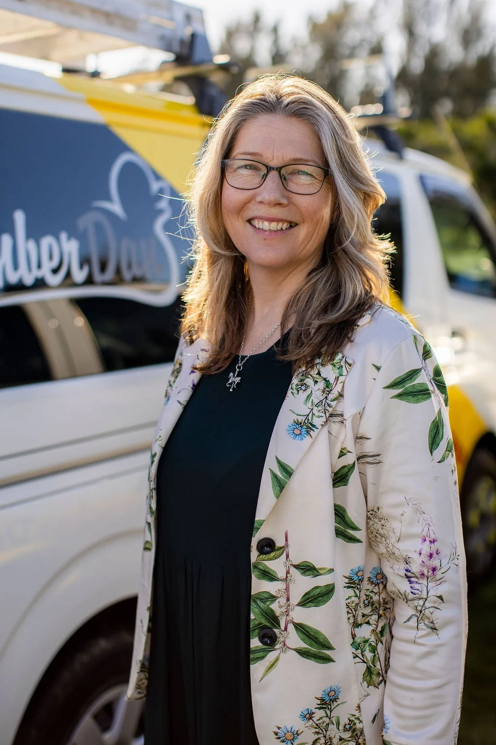 A woman with glasses and long, wavy hair is smiling outdoors, wearing a floral blazer over a black dress, standing in front of a vehicle with a sign that reads 'Super Duper Deli'.