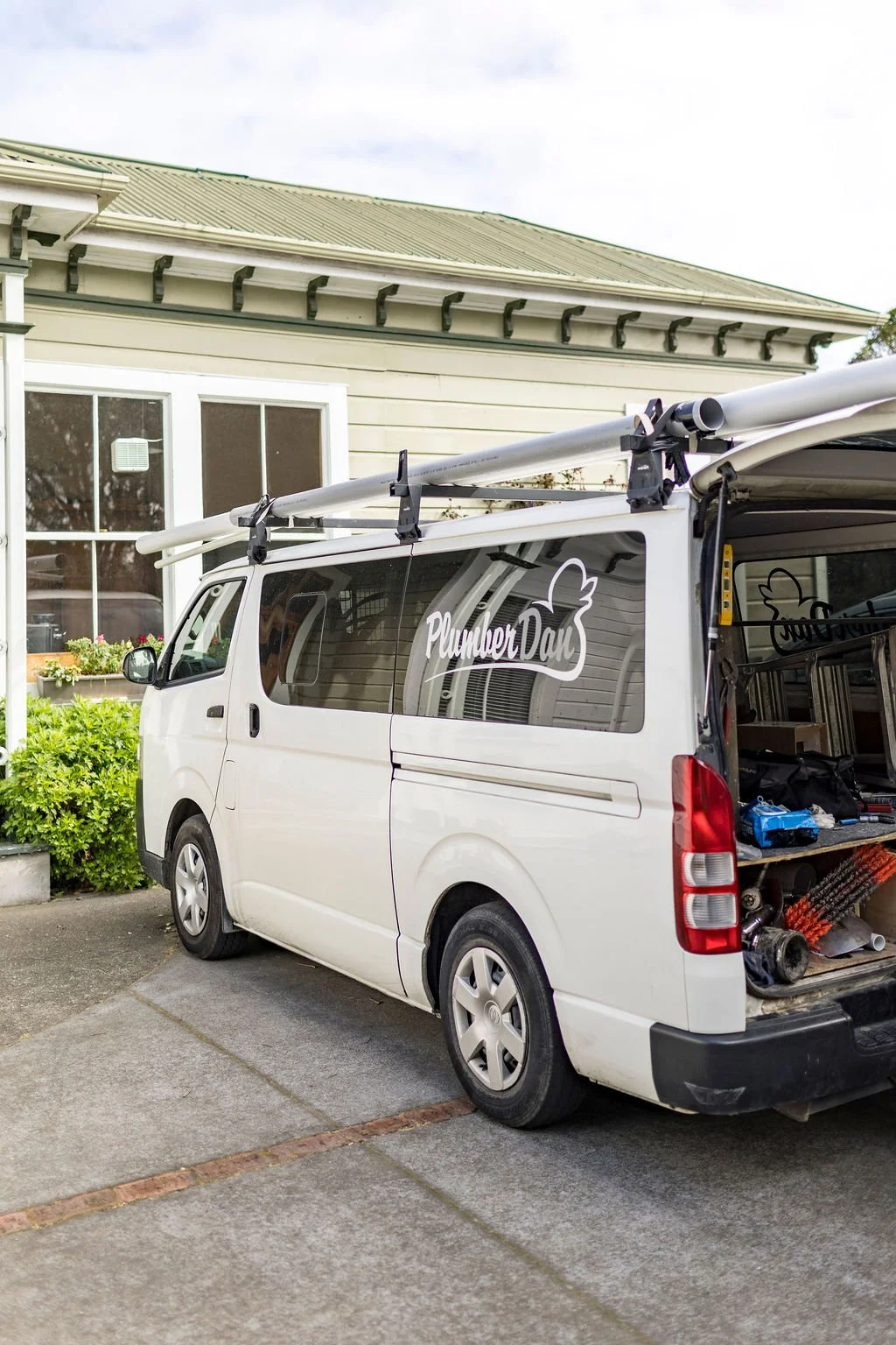 White service van parked outside a house, with a ladder on the roof and tools inside the back, featuring a 'Plumber Dave' logo.
