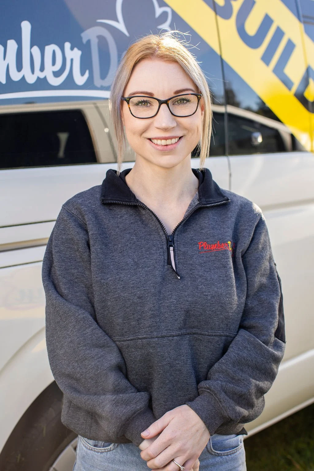 A woman with blonde hair, glasses, and a gray sweatshirt standing outdoors in front of a white vehicle with advertising signs.