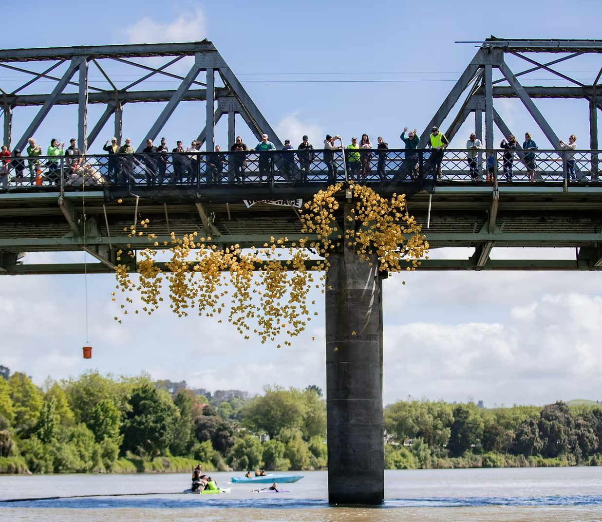 A bridge with people walking on it, with yellow balloons falling from beneath. Below, people are kayaking and paddleboarding on a river, with trees and a cloudy sky in the background.