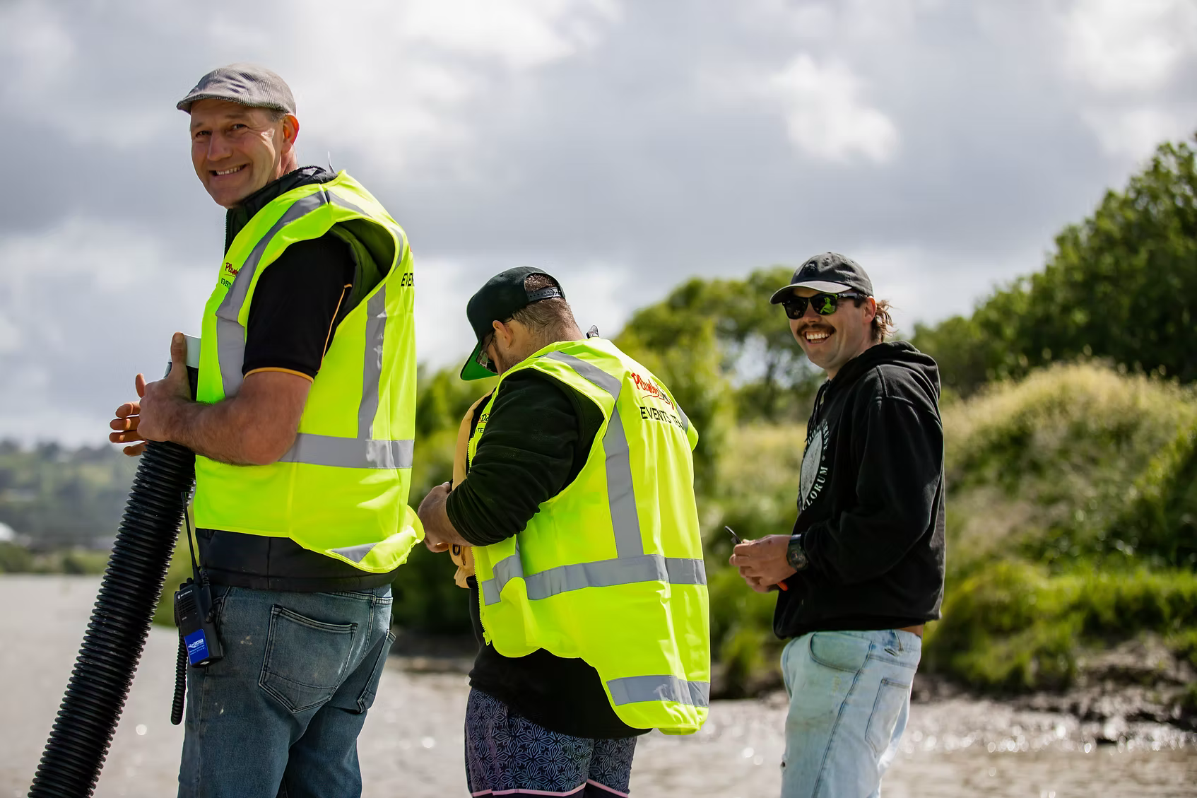 Three men standing outdoors near water, two wearing yellow reflective safety vests and one in a black hoodie with sunglasses, smiling and looking at each other.