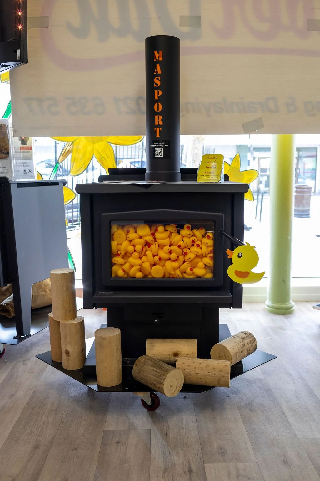 Duck-shaped rubber duckies in a heated display case with wooden logs in front, a black object marked 'MASSPORT' behind, and decorative yellow flowers painted on the window in the background.
