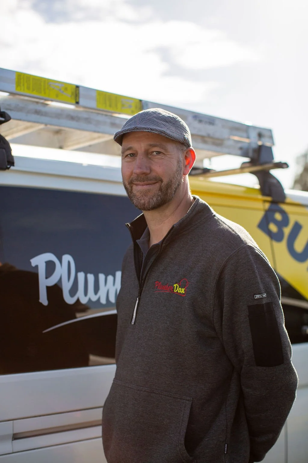 A man with a beard and gray flat cap standing outdoors near a white vehicle with a ladder on top and a sign that says "Plumber Dad."