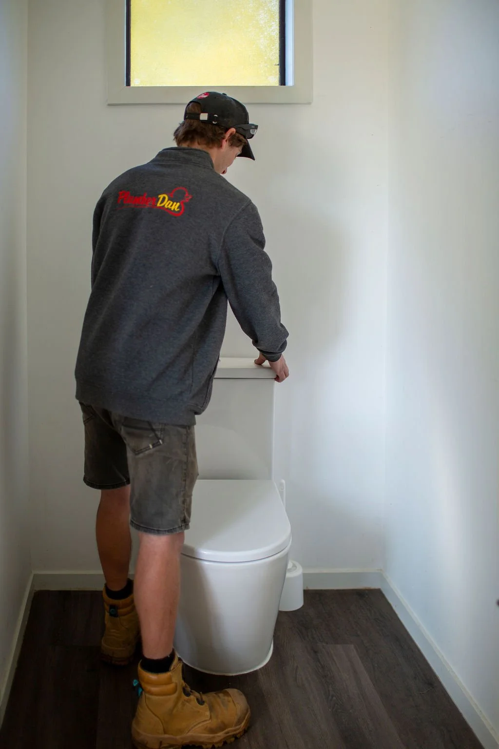 A man wearing a black cap, gray jacket with red and yellow logo on the back, and tan work boots, standing in front of a toilet in a small, plain bathroom with white walls and a window.