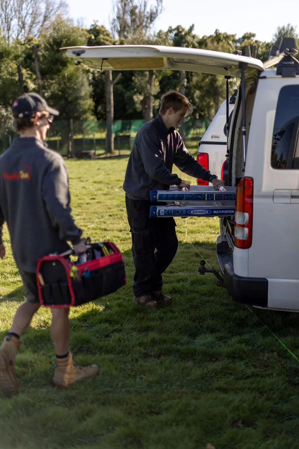Two men are setting up equipment from a white van outdoors on a grassy area. One man is wearing a gray jacket and cap, carrying a red toolbox, while the other is in a black jacket, handling equipment labeled 'Werner Multi-Position'.
