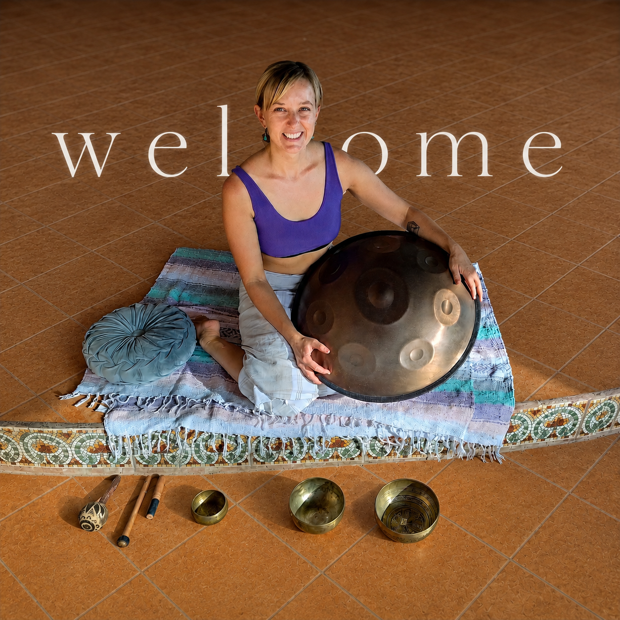 Woman sitting on a colorful cloth mat on a brown tiled floor, holding a large handpan instrument, with a round cushion beside her, three small metallic bowls, a wooden mallet, and two drumsticks arranged on the floor nearby, and a large word 'welcome' overlaid on the image.