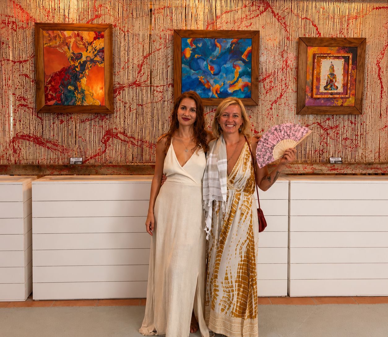 Two women standing in an art gallery, smiling at the camera. One woman is holding a pink fan. Behind them are three vibrant abstract paintings framed on a textured wall.