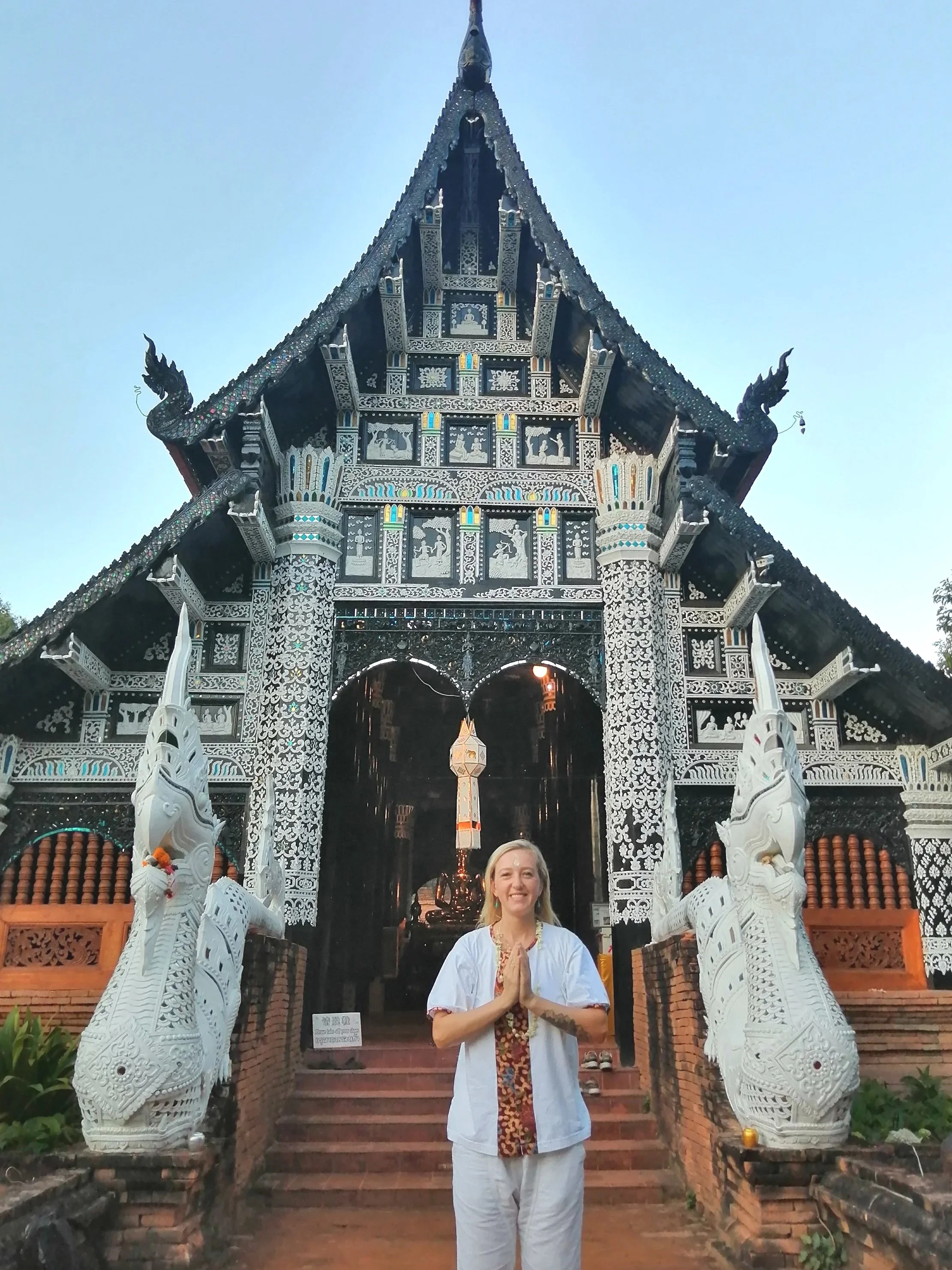 A woman in traditional white clothing and prayer posture stands at the entrance of a detailed, ornate temple with intricate carvings and sculptures, including two white serpent-like statues flanking the staircase.