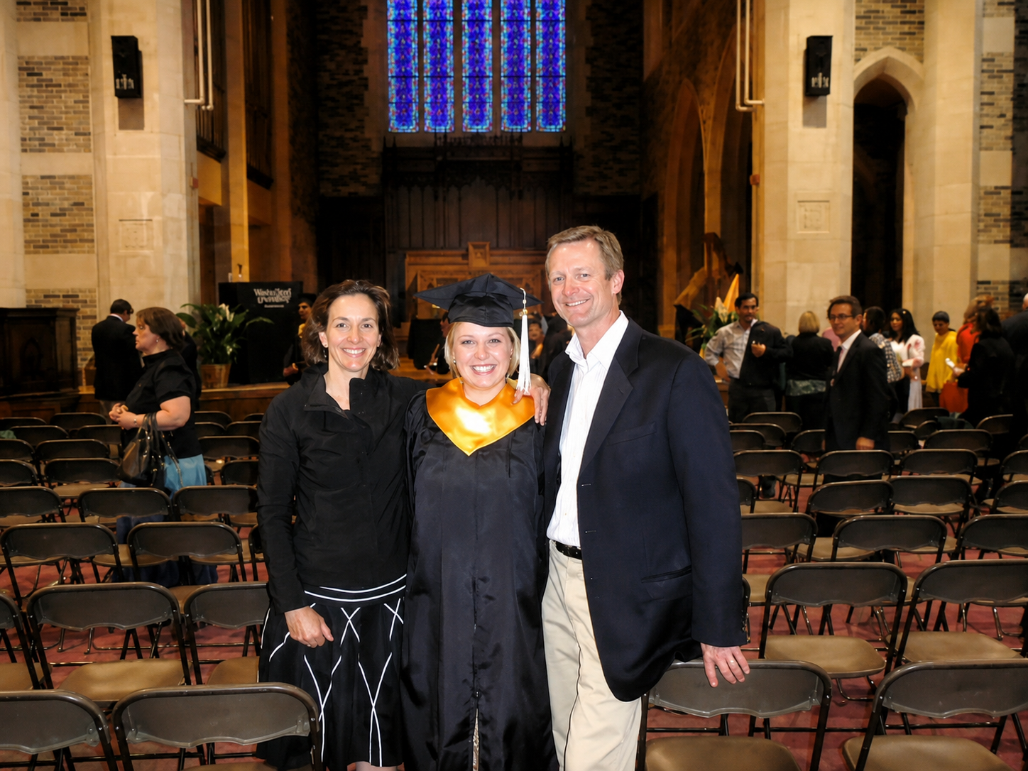 A woman in a graduation cap and gown with a gold stole, flanked by two people in formal attire, at a graduation ceremony in a large hall with stained glass windows and chairs.