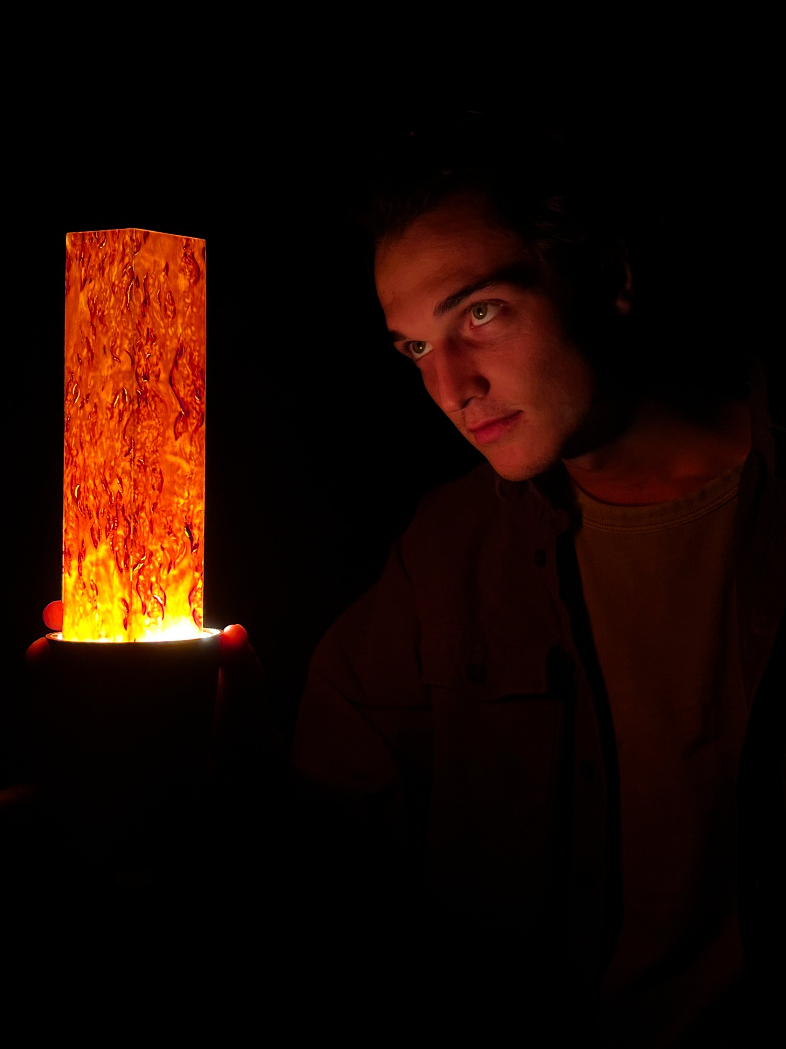 A young man in dim lighting holding a wood lamp prototype that glows in the dark.