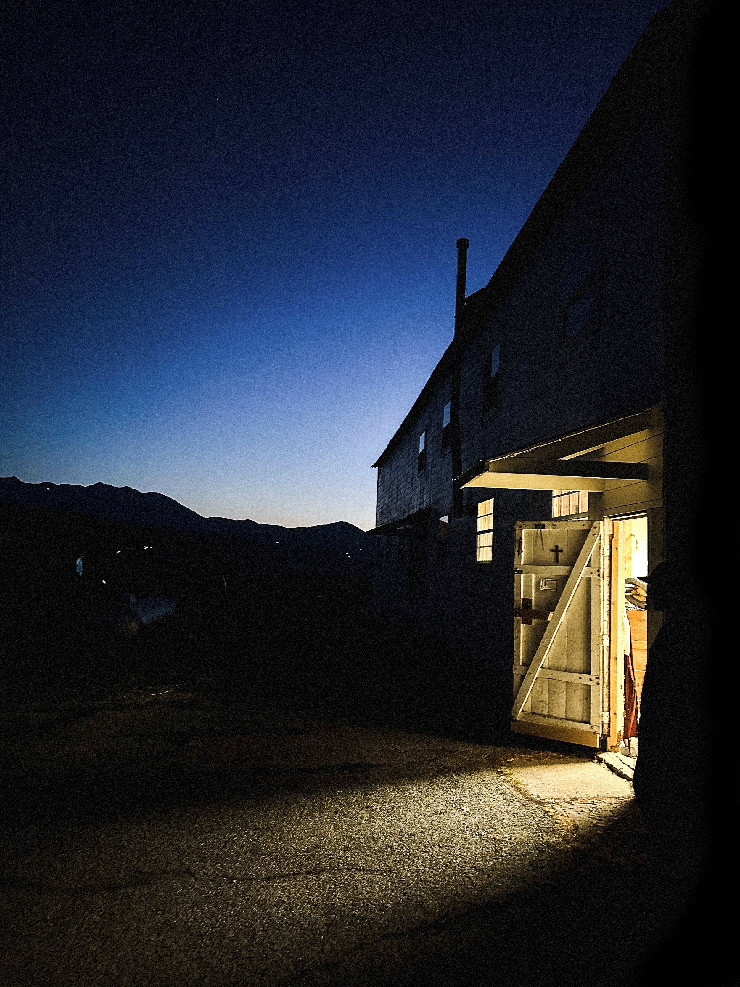 A building with a partially open door emitting light at dusk or dawn, with a mountain range in the background and a dark, textured ground in the foreground.