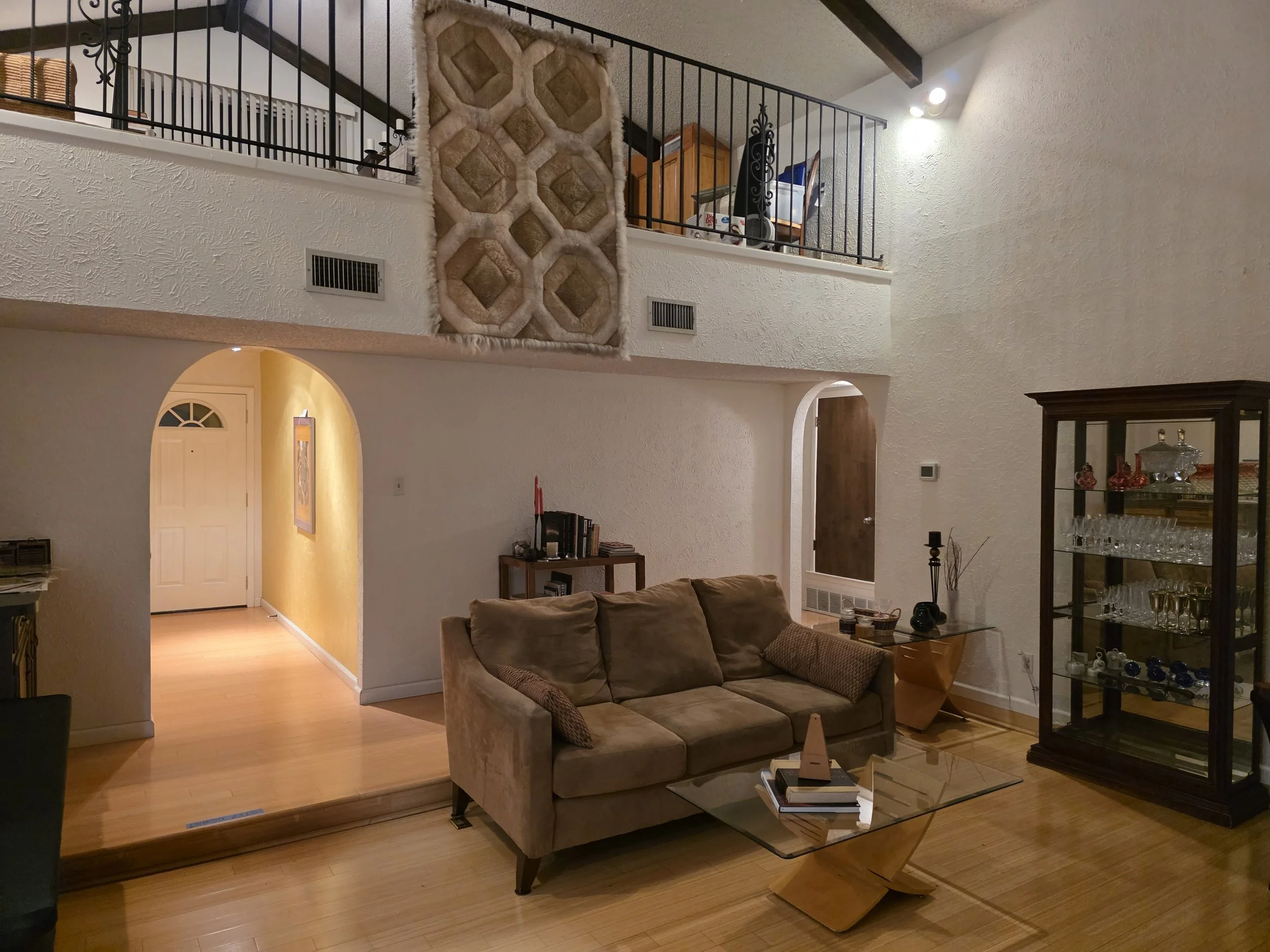 Living room with beige couch, glass coffee table, wood flooring, decorative cabinet with glassware, staircase, and a wall-mounted display.