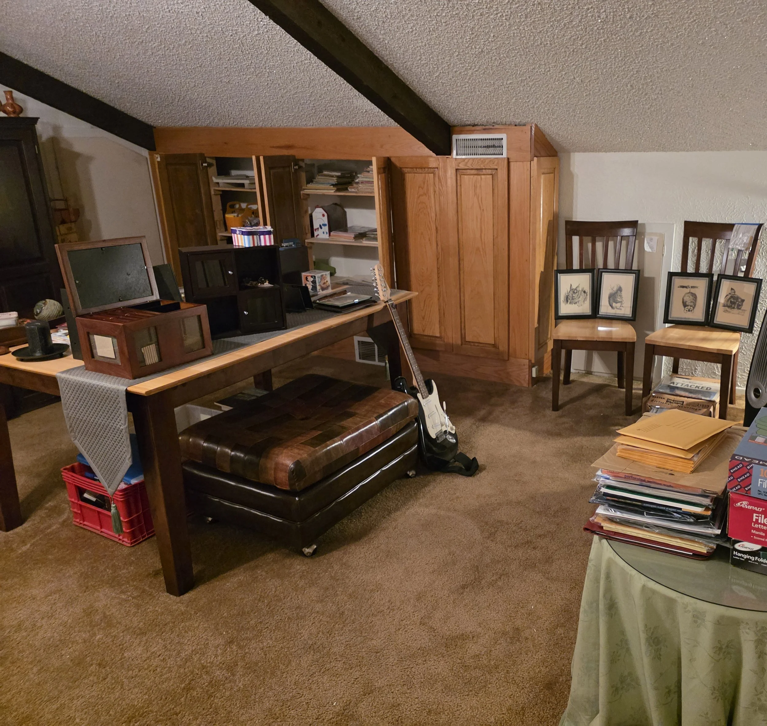 A room with a table, stacked papers, and framed drawings, with a wooden cabinet and chairs.