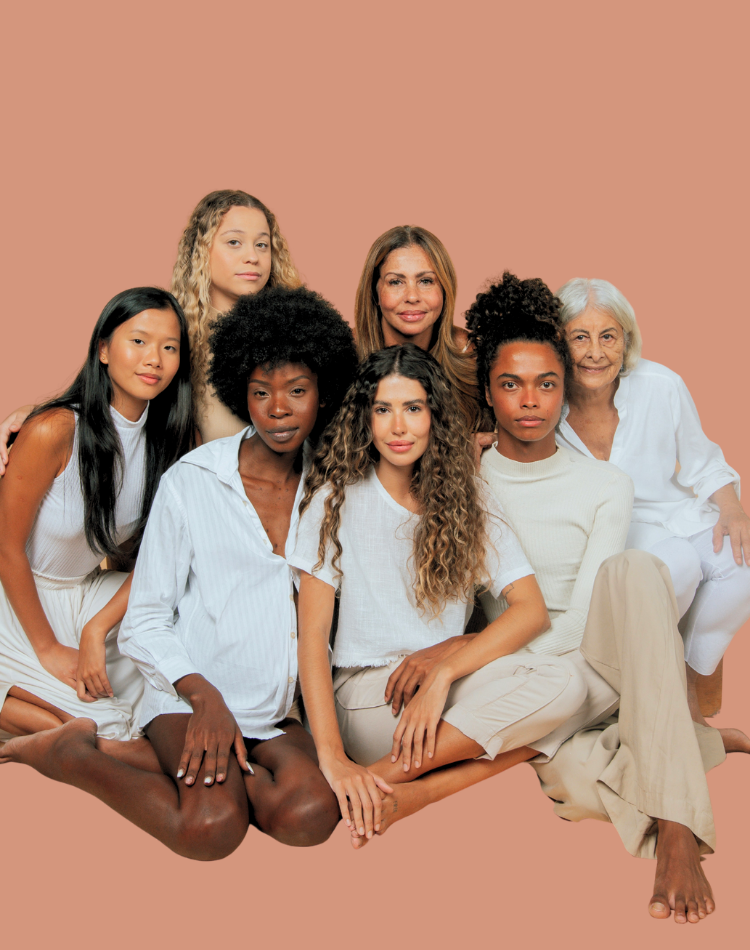 Seven women of varying ethnic backgrounds sitting together against a peach background, looking at the camera.