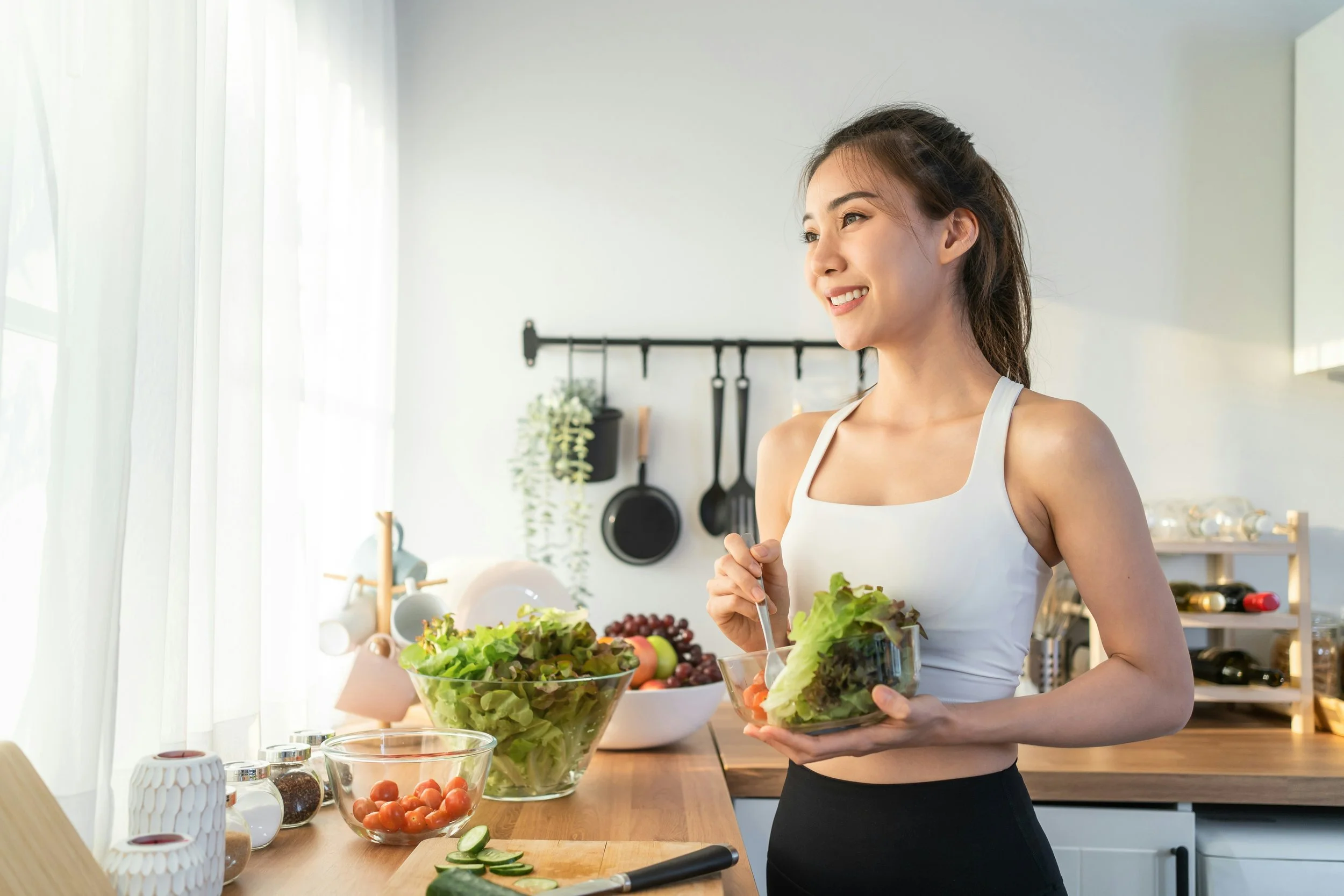 A woman in a white tank top preparing a salad in a modern kitchen with natural light. She holds a glass bowl of salad vegetables, with additional salad ingredients like cherry tomatoes, cucumbers, and leafy greens on the counter. The background shows kitchen utensils, a window with curtains, and a shelf with jars and plants.