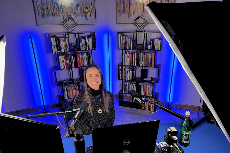 A woman with long brown hair wearing a black and gray sweater sitting at a desk with a computer and microphone, in a room with blue LED lights, bookshelves, and wall art.