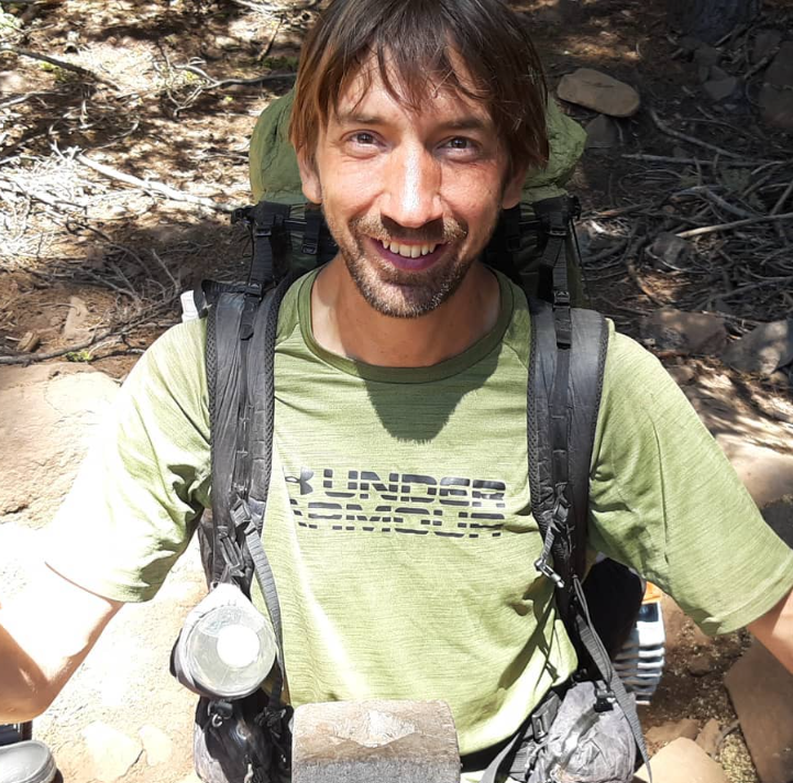 A man hiking in a forest, smiling, wearing a green Under Armour shirt, carrying a backpack with gear attached, surrounded by trees and rocks.