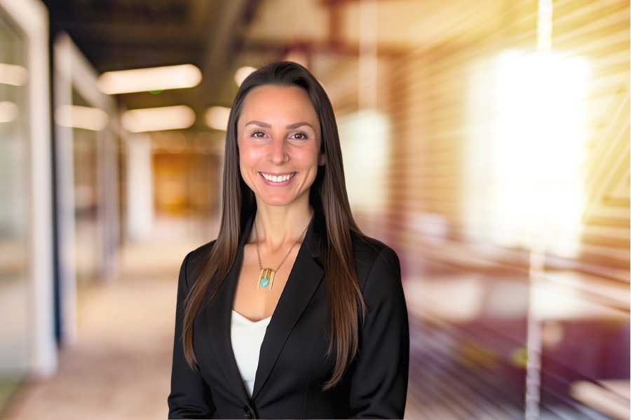 A smiling woman in professional attire standing in a modern office corridor with warm sunlight streaming in.