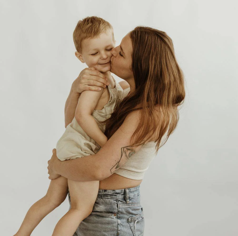 A woman with red hair and a tattoo on her arm is lifting a young boy, who has red hair, and giving him a kiss on the cheek. The boy is smiling, and they are standing against a plain white background.