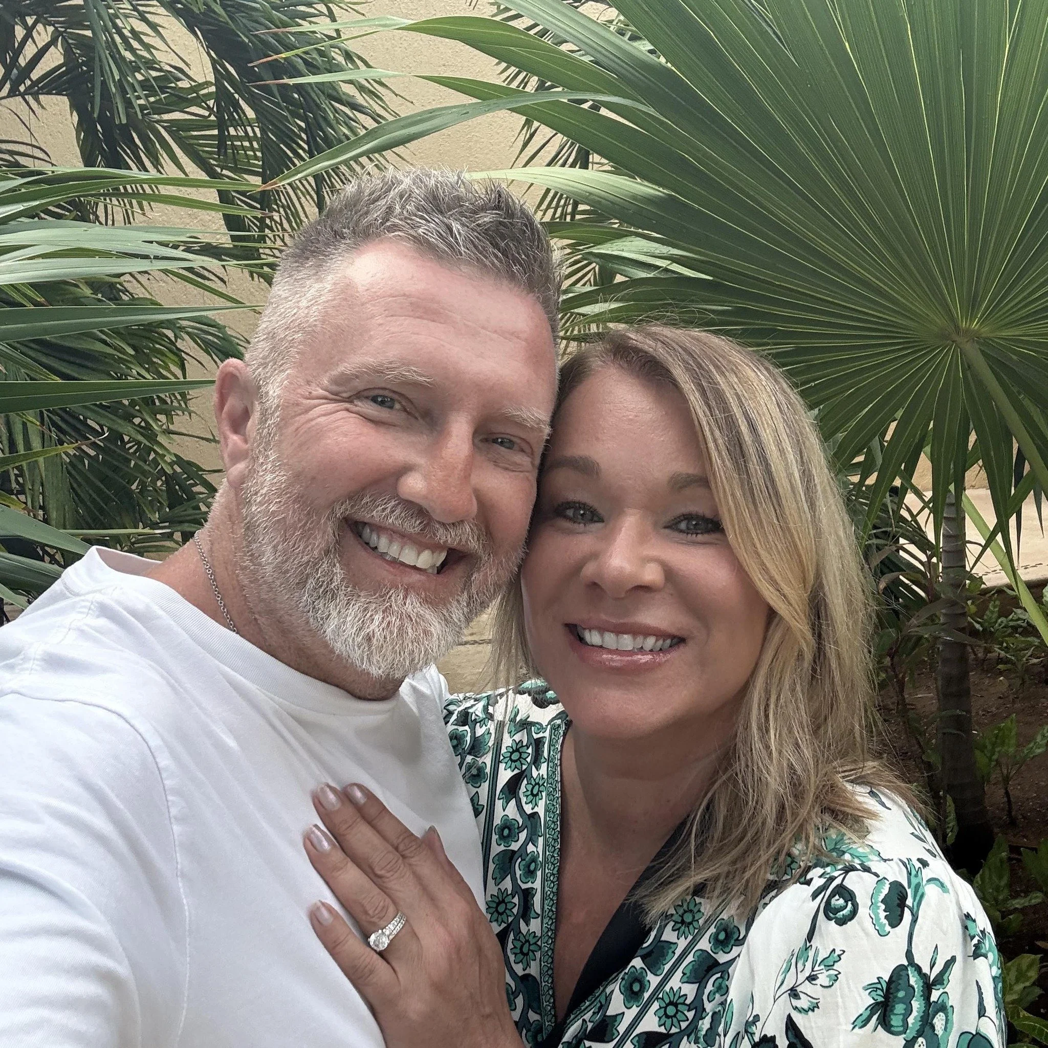 A close-up photo of a middle-aged man with gray hair and a beard, smiling, and a young woman with blonde hair, smiling, both standing in front of large green tropical plants.