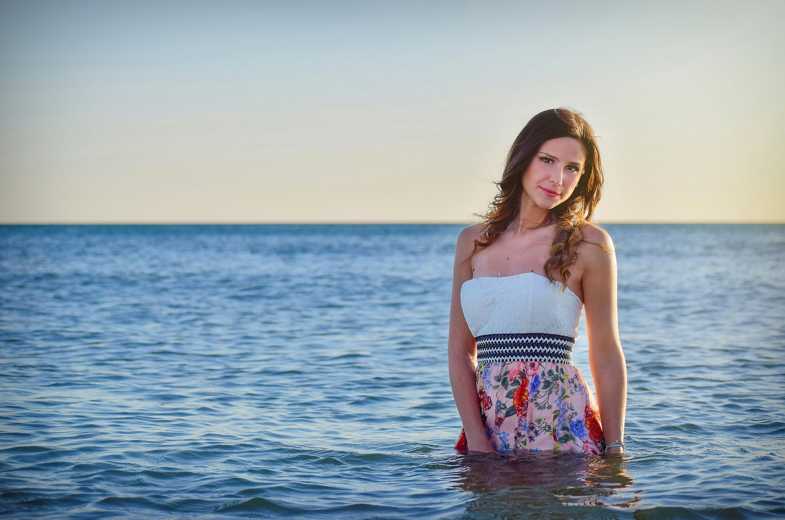 A young woman with brown hair wearing a strapless white top and a floral skirt, standing in the ocean with water up to her thighs during sunset, with a calm sea and a distant horizon in the background.