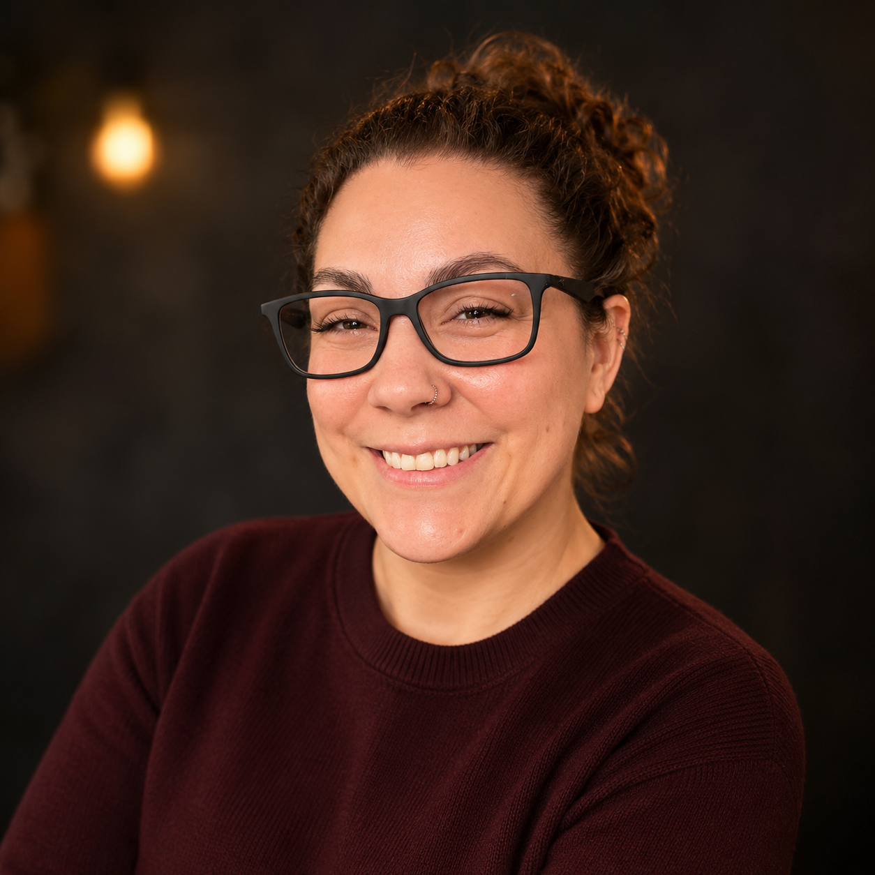 A woman with curly brown hair pulled back, wearing black glasses, smiling, wearing a dark red top, in front of a dark background with warm lighting.