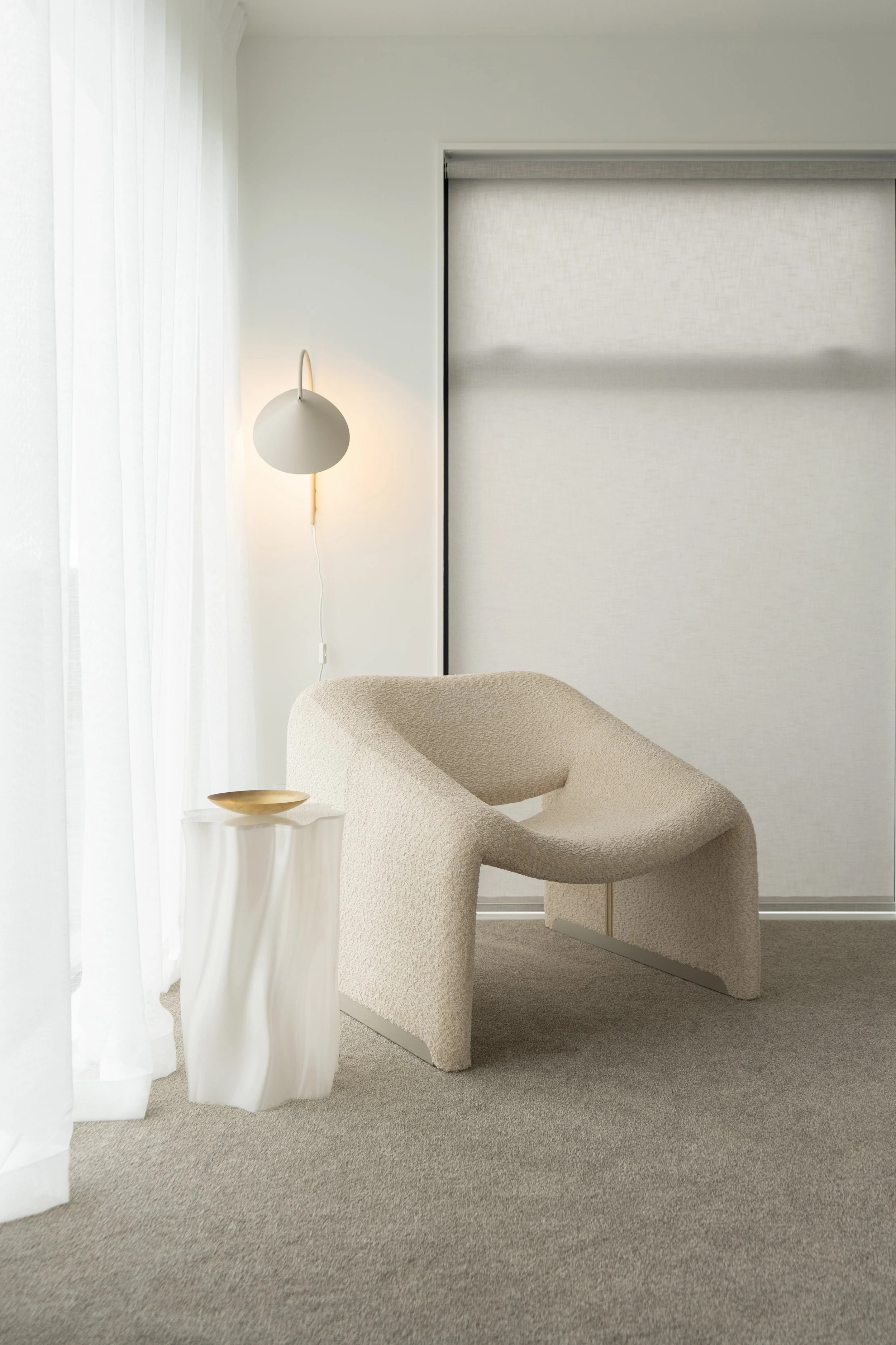 Minimalist living space with a beige armchair, a white column side table with a gold bowl, sheer white curtains, a white wall-mounted lamp, and a textured white sliding door.