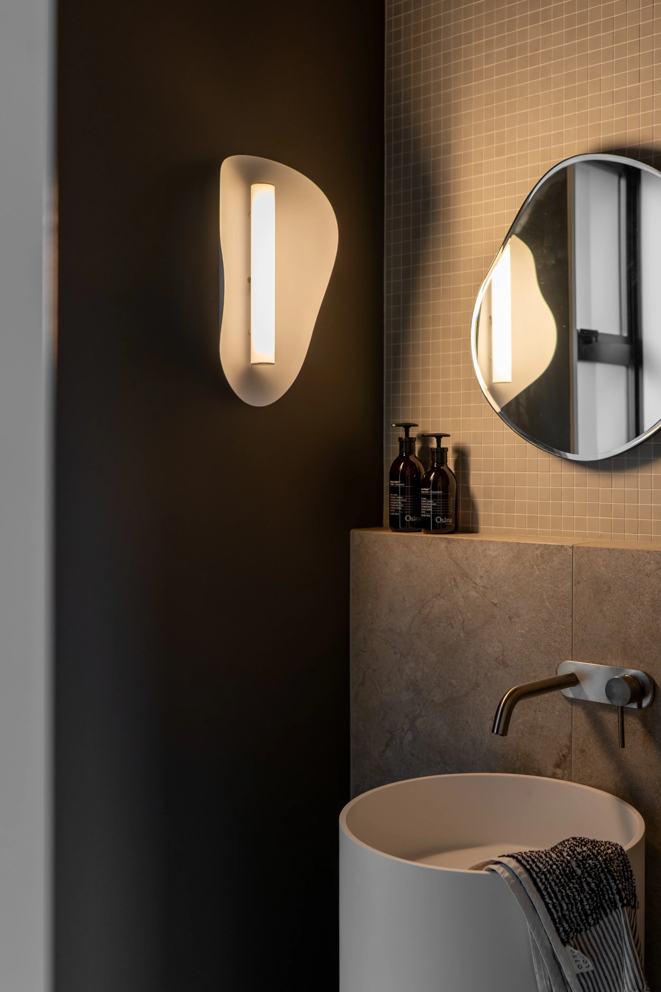 Modern bathroom with a dark wall-mounted light fixture, a mirror, toiletries, a faucet, and a white sink with a towel draped over it.